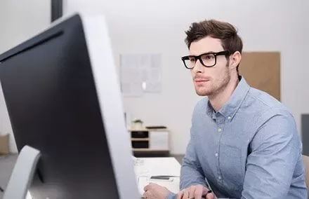 Homem de óculos trabalha em computador em escritório.