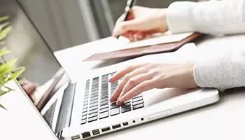 Pessoa digitando em um laptop prateado e escrevendo em um caderno.