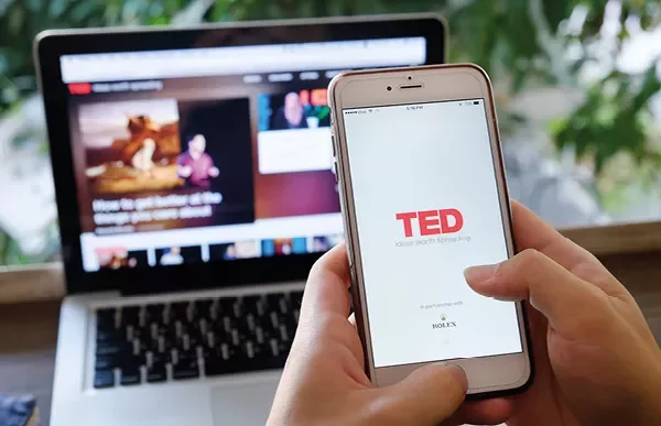Pessoa segurando um celular com o aplicativo TED aberto, e um laptop ao fundo.